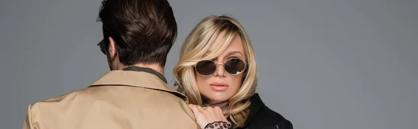 Blonde in stylish sunglasses hugging man in beige jacket isolated on grey, banner — Stockfoto