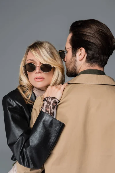 Blonde in stylish sunglasses hugging bearded man in beige jacket isolated on grey — Stock Photo