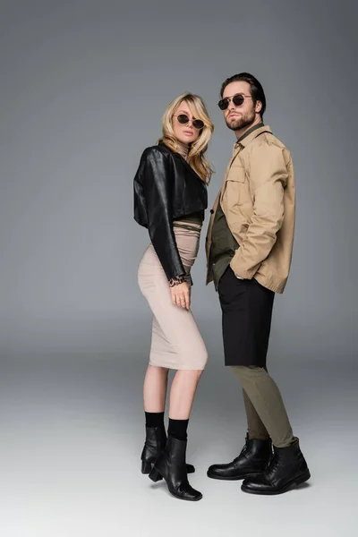 Full length of stylish woman in autumnal outfit and sunglasses posing near man standing with hand in pocket on grey — Stockfoto