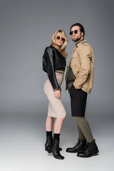 Full length of stylish woman in autumnal outfit and sunglasses posing near man standing with hand in pocket on grey — Photo de stock