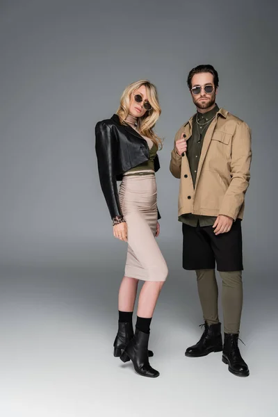 Full length of stylish couple in autumnal outfits and sunglasses posing and looking at camera on grey — Stock Photo