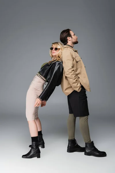 Full length of stylish woman in trendy outfit and sunglasses leaning on man while posing on grey — Stock Photo