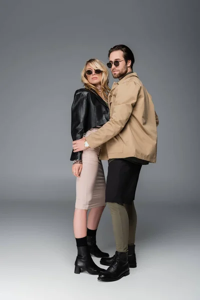 Full length of stylish man in autumnal outfit and sunglasses posing with woman on grey — Stockfoto