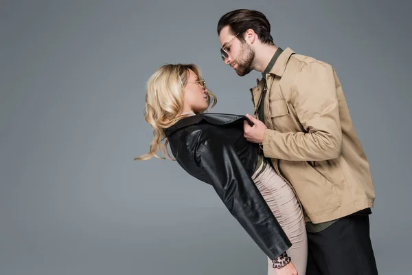 Side view of bearded man in sunglasses pulling leather jacket of blonde woman while flirting isolated on grey — Foto stock