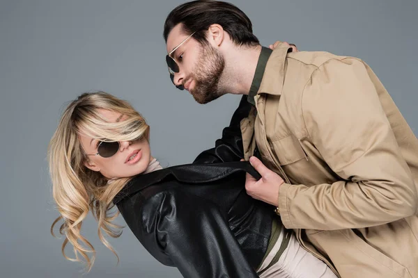Bearded man in sunglasses pulling leather jacket of blonde woman while flirting isolated on grey - foto de stock