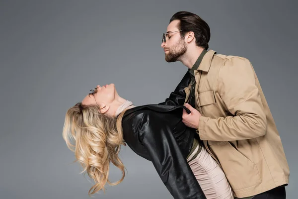 Side view of stylish man in sunglasses pulling leather jacket of blonde woman isolated on grey - foto de stock