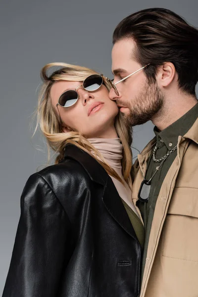 Бородатый мужчина целует щеку блондинки и стильной женщины в солнечных очках, изолированных на сером — стоковое фото