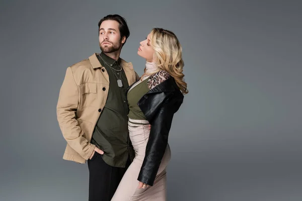 Blonde woman in black leather jacket looking at man posing with hand in pocket isolated on grey — Stock Photo