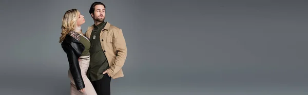 Блондинка в черной кожаной куртке смотрит на мужчину, позирующего с рукой в кармане, изолированного на сером, баннер — стоковое фото