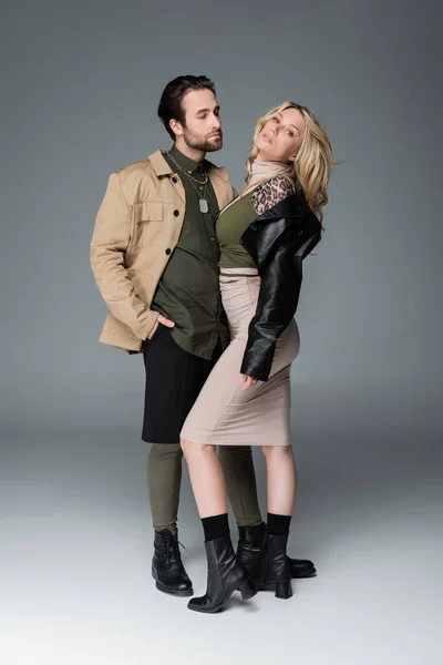 Full length of stylish man in autumnal outfit posing with hand in pocket near blonde woman on grey — Stock Photo