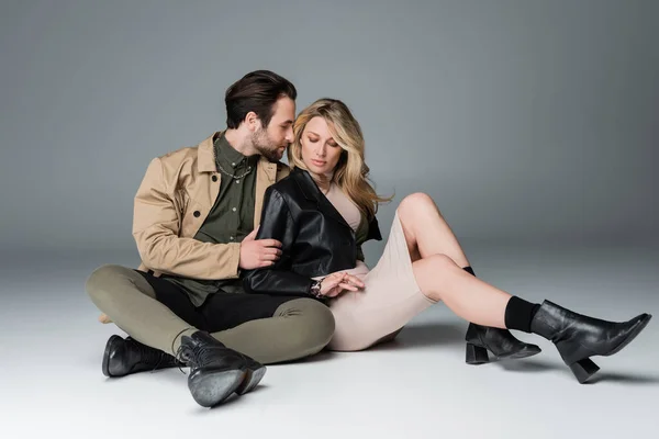 Stylish bearded man and blonde woman in trendy outfits sitting in black boots on grey — Photo de stock