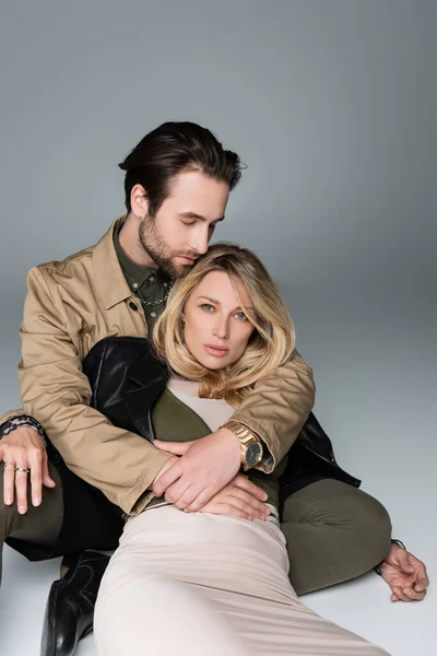 Stylish bearded man hugging blonde woman in trendy outfit sitting and looking at camera on grey — Foto stock