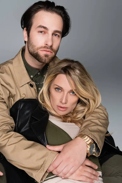 Bearded man hugging blonde woman in trendy outfit and looking at camera on grey — Stock Photo