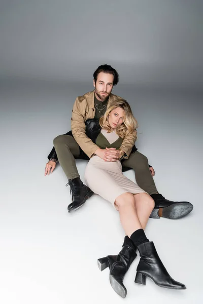 Full length of stylish bearded man and blonde woman in trendy outfits sitting while looking at camera on grey — Fotografia de Stock