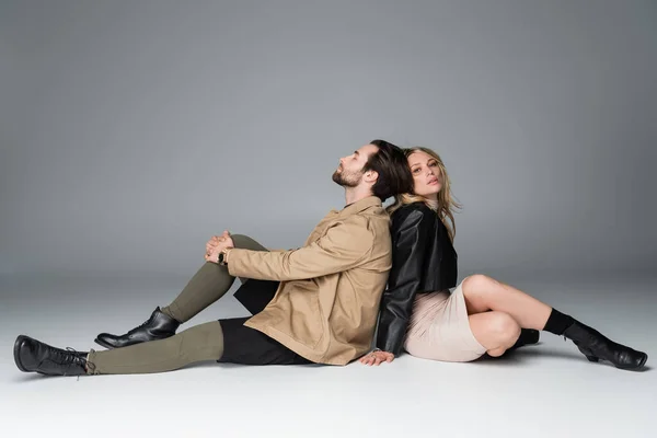 Full length of stylish bearded man and blonde woman in trendy outfits sitting back to back on grey - foto de stock