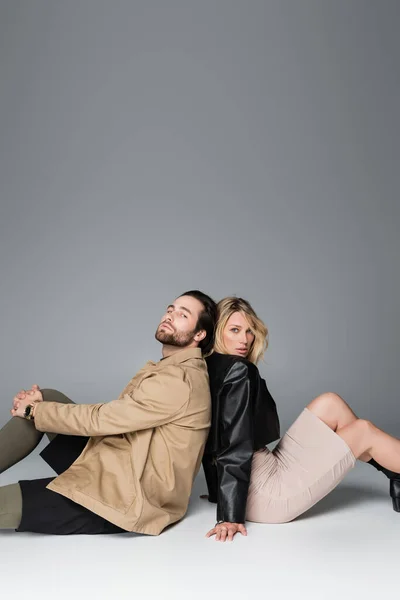 Bearded man and blonde woman in stylish outfits sitting back to back on grey — Photo de stock