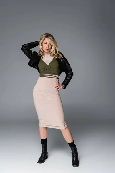 Full length of blonde woman in beige dress and black leather jacket posing with hand on hip on grey — Photo de stock
