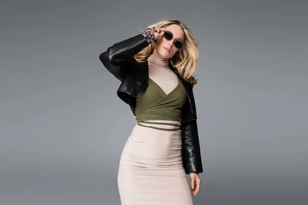 Блондинка в черной кожаной куртке регулирует модные солнцезащитные очки, изолированные на сером — стоковое фото