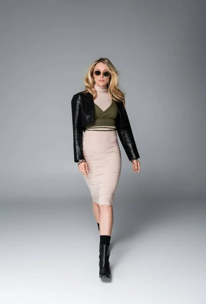 Full length of blonde woman in black leather jacket and stylish sunglasses walking on grey — Stock Photo