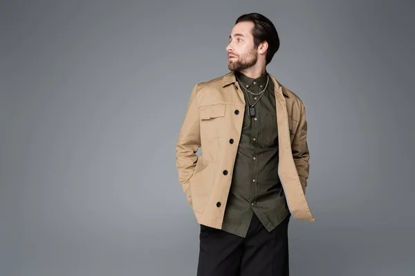 Bearded man in trendy outfit posing with hands in pockets and looking away isolated on grey — Stock Photo