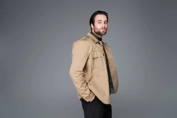 Bearded man in beige jacket posing with hands in pockets and looking at camera isolated on grey — Stockfoto