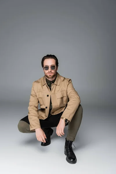 Full length of bearded man in stylish outfit and sunglasses sitting while posing on grey — Stock Photo