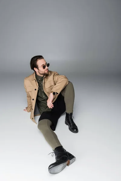 Full length of bearded man in stylish outfit and sunglasses sitting on grey - foto de stock