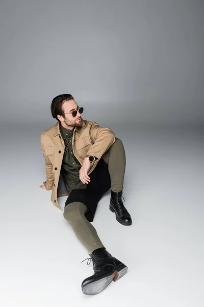 Full length of bearded man in stylish outfit and sunglasses sitting on grey — Stock Photo