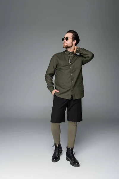 Полная длина бородатый человек в солнцезащитных очках и стильный наряд позируя с рукой в кармане на сером — стоковое фото