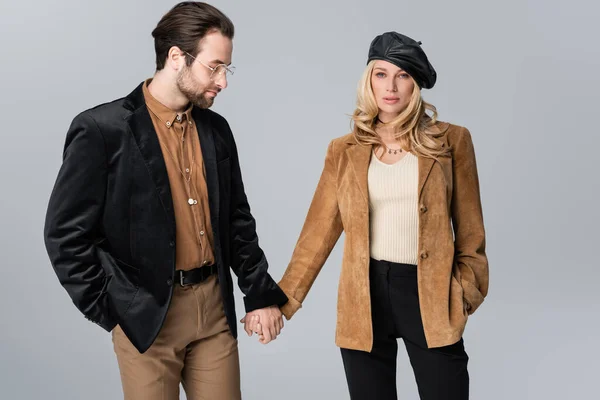 Blonde woman in stylish beret holding hands with bearded man in sunglasses isolated on grey — Foto stock