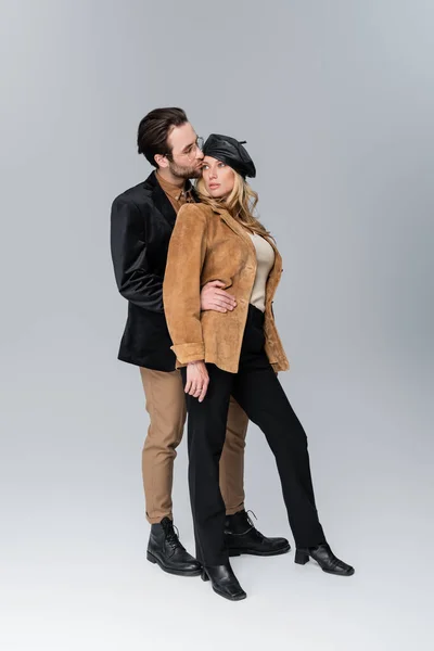 Full length of bearded man in sunglasses kissing blonde woman in stylish beret on grey — Stockfoto