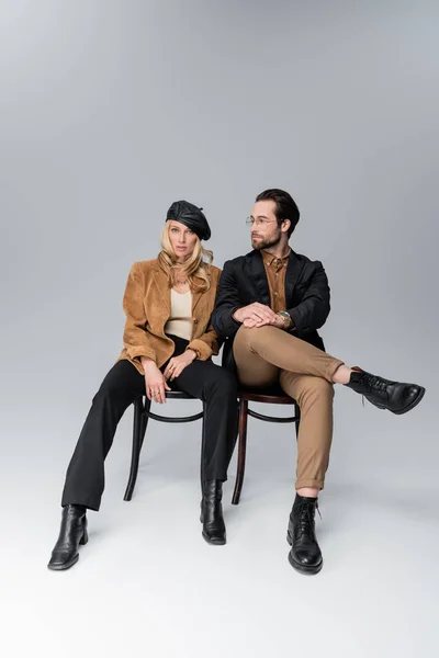 Full length of blonde woman in stylish beret near bearded man in sunglasses sitting on chairs on grey — Photo de stock