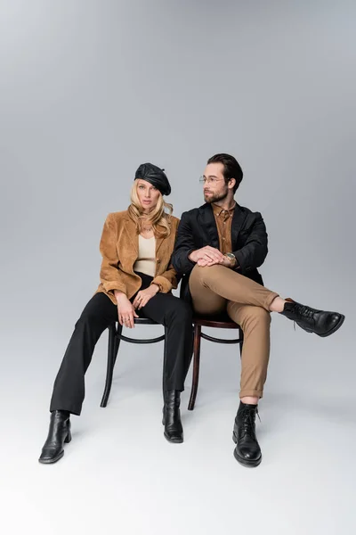 Full length of blonde woman in stylish beret near bearded man in sunglasses sitting on chairs on grey — Fotografia de Stock