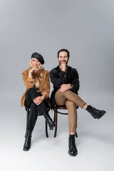 Full length of happy blonde woman in stylish beret near bearded man in sunglasses sitting on chairs on grey — Photo de stock