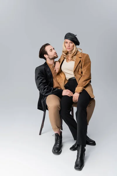 Full length of blonde woman in stylish beret sitting on knees of bearded man in sunglasses on grey — Stock Photo