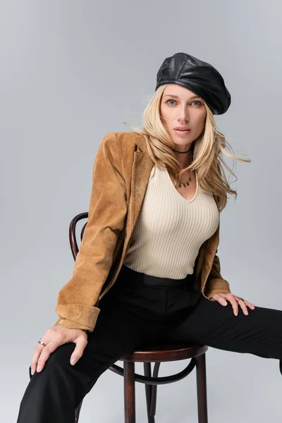 Blonde model in stylish leather beret and beige blazer sitting on chair isolated on grey - foto de stock