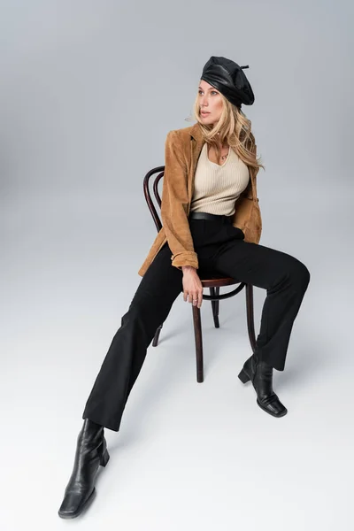 Full length of blonde woman in stylish leather beret and beige blazer posing while sitting on chair on grey - foto de stock