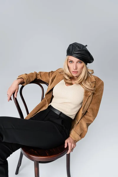 Blonde woman in stylish leather beret and beige blazer sitting on chair isolated on grey — Fotografia de Stock