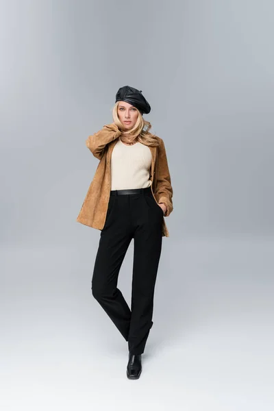 Blonde woman in stylish leather beret and beige blazer posing with hand in pocket on grey — Foto stock