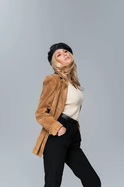 Блондинка в стильном кожаном берете и бежевом пиджаке позирует с руками в карманах, изолированных на сером — стоковое фото