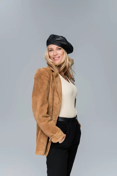 Happy blonde woman in stylish leather beret and beige blazer posing with hands in pockets isolated on grey — Stock Photo