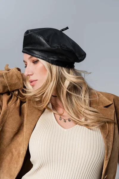 Блондинка в стильном кожаном берете и бежевом пиджаке позирует изолированно на сером — стоковое фото