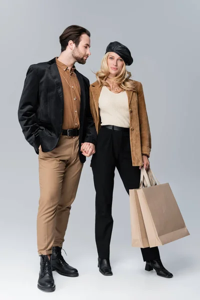 Bearded man holding hands with blonde woman in stylish beret standing with shopping bags on grey — Photo de stock