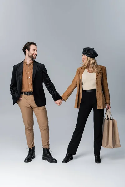 Happy man holding hands with blonde woman in stylish beret standing with shopping bags on grey — Foto stock