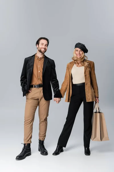 Cheerful man holding hands with blonde woman in stylish beret standing with shopping bags on grey — Stock Photo