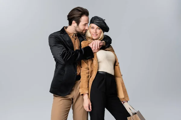 Bearded man hugging happy blonde woman in stylish beret holding shopping bags isolated on grey — Stock Photo