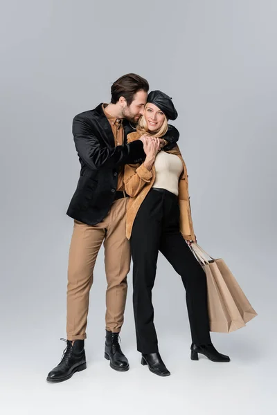 Bearded man hugging happy woman in stylish beret holding shopping bags on grey — Photo de stock