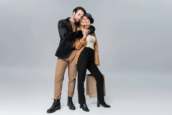 Cheerful man hugging happy woman in stylish beret holding shopping bags on grey — Stockfoto