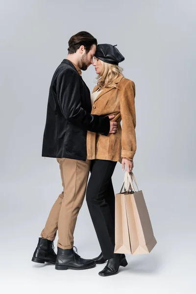 Side view of bearded man hugging blonde girlfriend in stylish beret holding shopping bags on grey — Fotografia de Stock