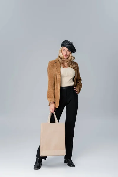 Full length of blonde woman in stylish leather beret and beige blazer holding shopping bag on grey — Stockfoto